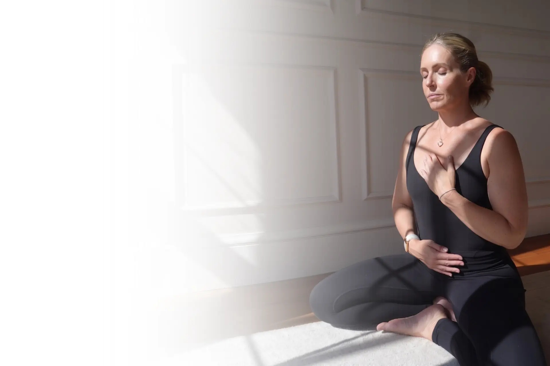 Jen seated in meditation, one hand on her chest and one on her belly, in warm natural light